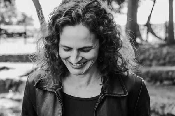 Black and White image of Jessica Cuello in leather jacket, smiling, walking through woodded area.